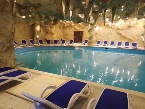 An indoor pool area is surrounded by natural stone walls and illuminated by soft lighting. Blue lounge chairs are arranged neatly around the pool, which reflects the overhead lights, creating a serene environment for relaxation.