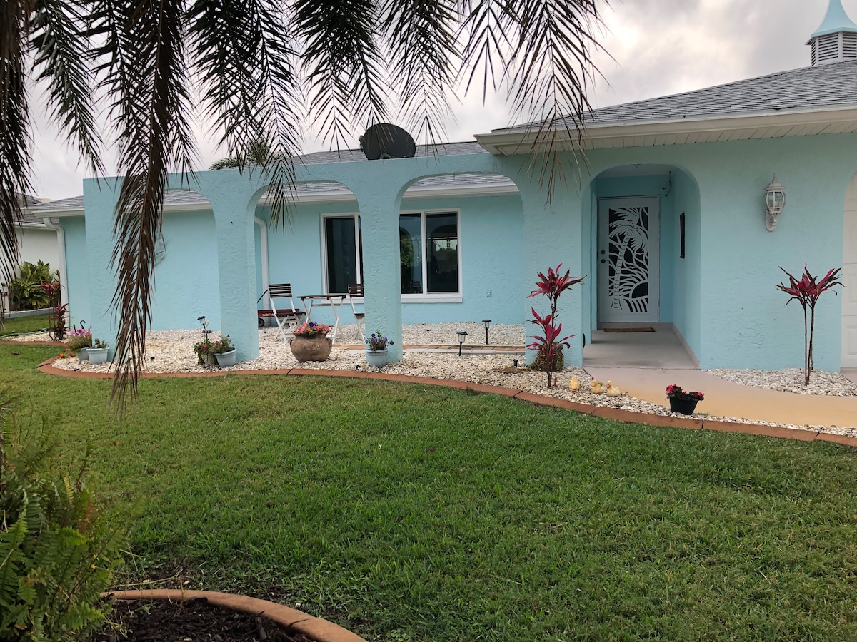 A light blue house is surrounded by a well-maintained lawn and decorative landscaping. Two chairs are positioned on a small patio area under an archway, with potted plants adding color to the scene. The entrance features a stylized door with intricate design details.