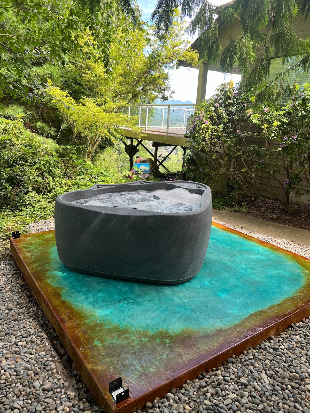 A hot tub is situated on a vibrant, turquoise platform surrounded by a gravel area and lush greenery. The backdrop features trees and a view of a wooden deck. Soft sunlight reaches the hot tub, enhancing the serene outdoor setting.