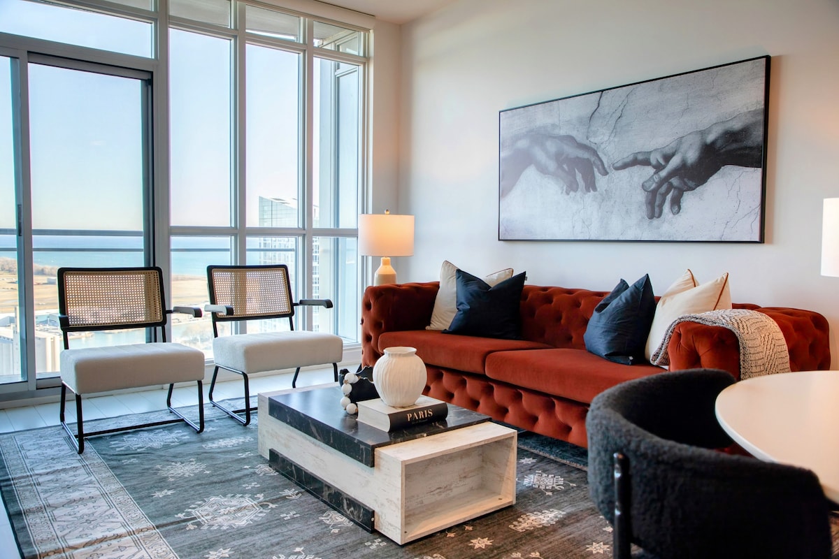 The living area features a striking red sofa adorned with decorative cushions, paired with two stylish armchairs. A modern coffee table sits centrally, showcasing decorative items. Large windows allow natural light to brighten the space and offer a view of the lake beyond.