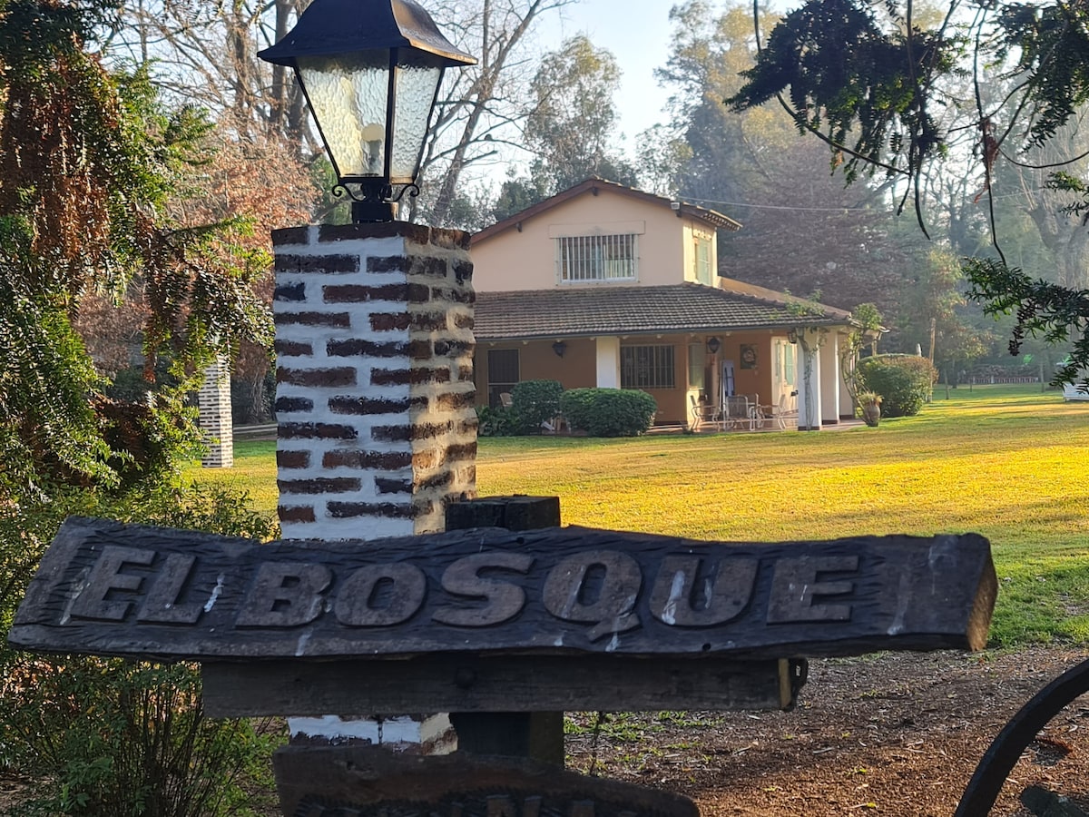A welcoming house is set among lush greenery, featuring a light-colored exterior and a spacious front porch. A wooden sign bearing the name 'El Bosque' is prominently displayed, accompanied by a lantern on a rustic stone post, conveying a serene environment.