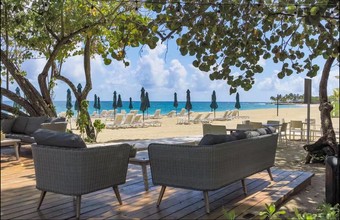 A shaded outdoor seating area features stylish wicker furniture, offering comfort with soft cushions. In the background, beach chairs and umbrellas are arranged along a sandy shore, with turquoise water extending to the horizon under a partly cloudy sky.
