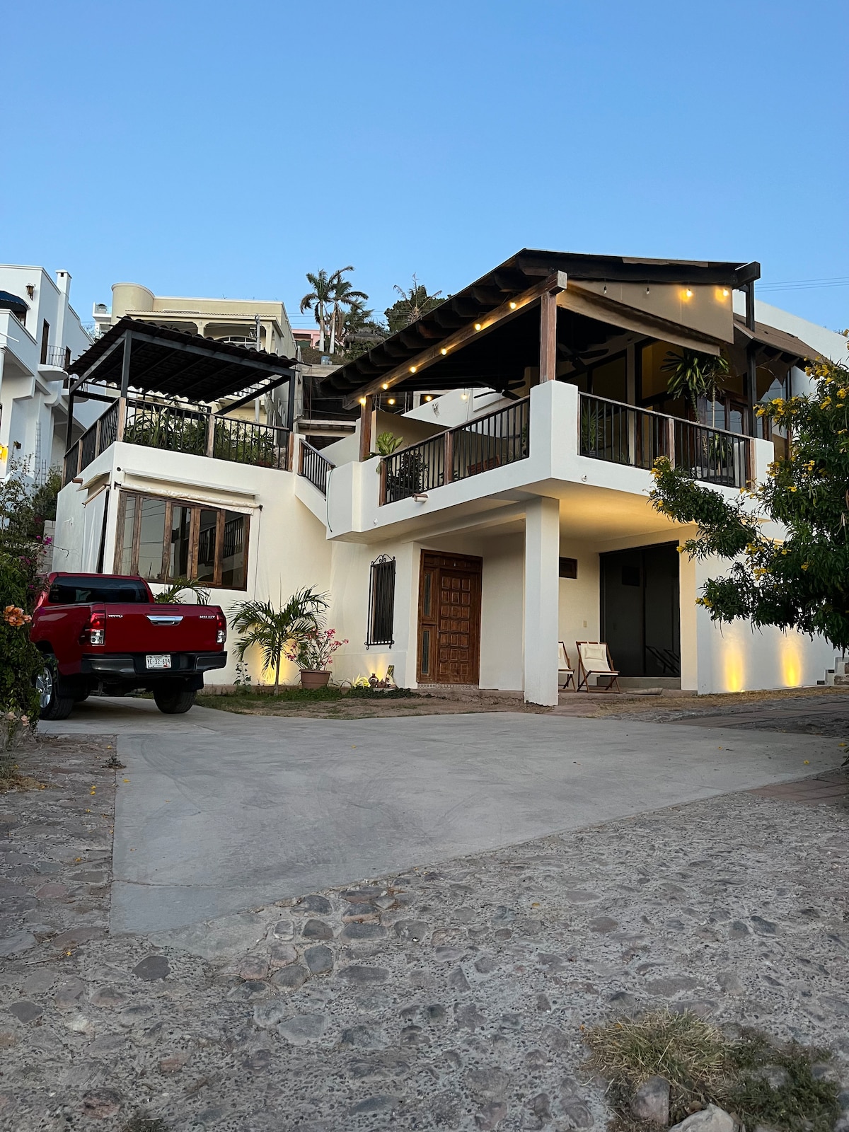 A two-story house is depicted, featuring a combination of white exterior walls and natural wood accents. Warm exterior lighting enhances the entrance, while a stone pathway leads to a spacious front lawn. A vehicle is parked in the driveway, surrounded by lush greenery.