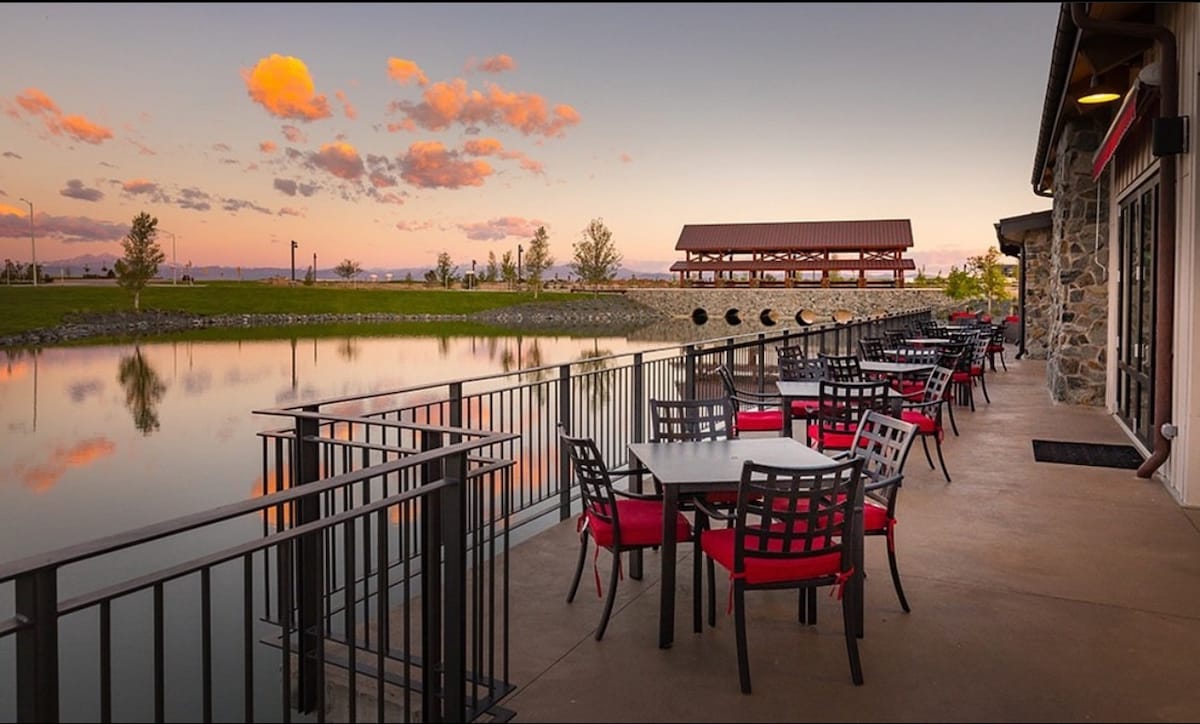 An outdoor dining area is depicted along a tranquil waterway, featuring black metal tables with red cushions. A picturesque view includes a covered pavilion across the water, with a backdrop of distant hills under a soft, colorful sunset.