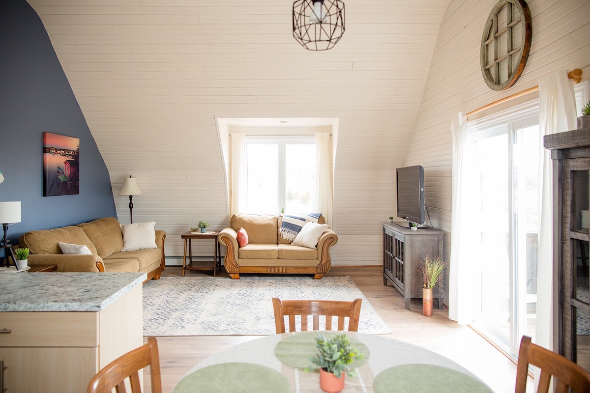 A sunlit living area features a comfortable couch and a cozy armchair, complemented by a small side table with a lamp. A television is situated next to the window, which offers views of the outdoors. A dining table is visible nearby, adorned with a small potted plant.