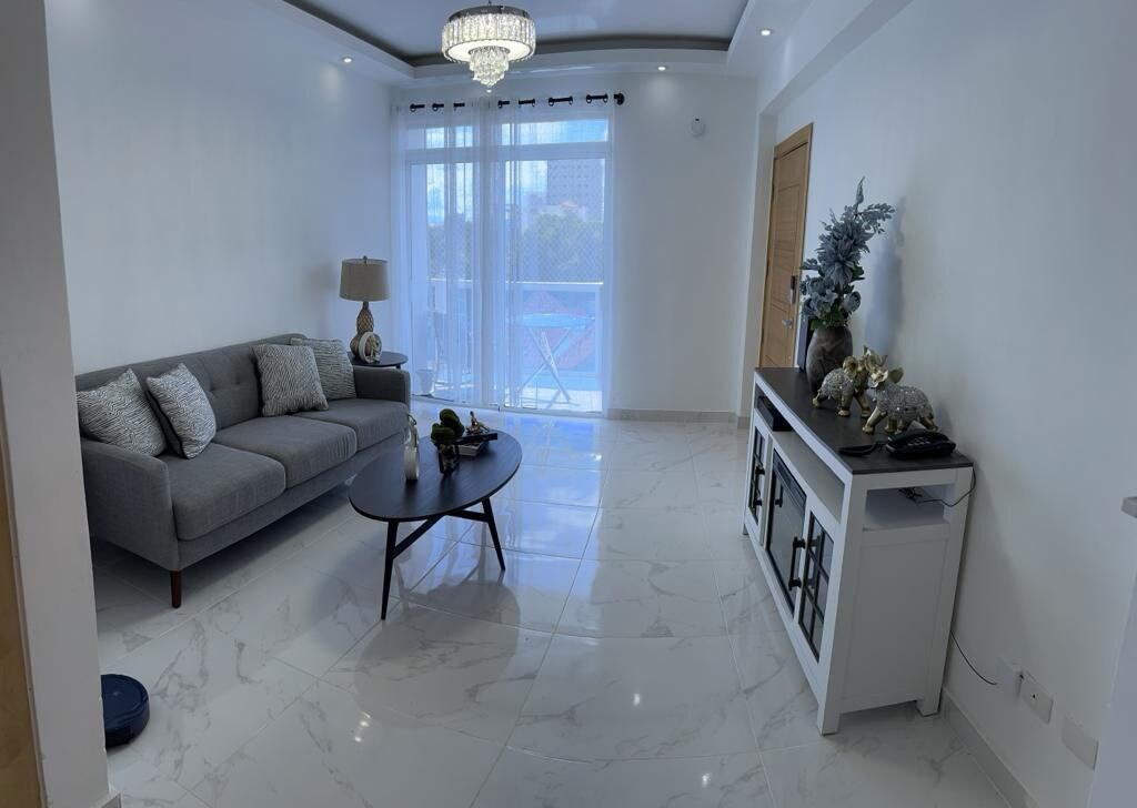 A bright living area features a comfortable gray sofa adorned with decorative pillows. A round coffee table sits in front, complemented by a sleek side console. The space is illuminated by natural light streaming through sheer curtains, with a balcony door offering scenic views.