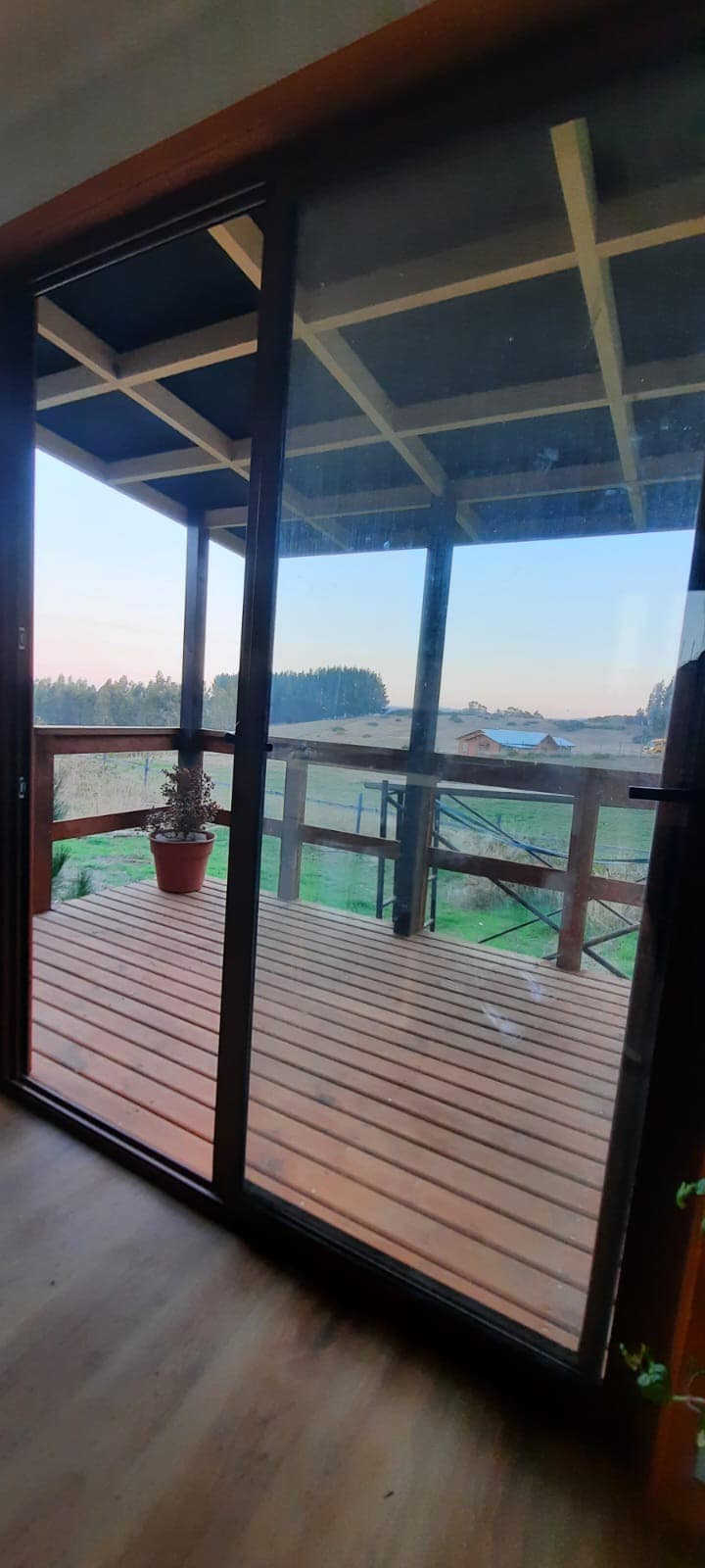 A sliding glass door opens to a wooden deck, bordered by a railing. A potted plant adds greenery to the space, while views of the surrounding rural landscape are visible beyond. The soft light of early morning or late afternoon can be seen illuminating the scene.