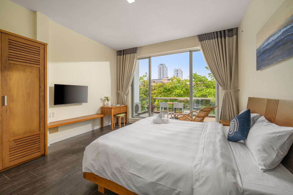 A bright bedroom features a spacious bed with a white comforter, complemented by a decorative pillow. Large windows allow natural light to fill the room, showcasing a view of greenery and nearby buildings. A wooden closet and a TV mounted on the wall are included.