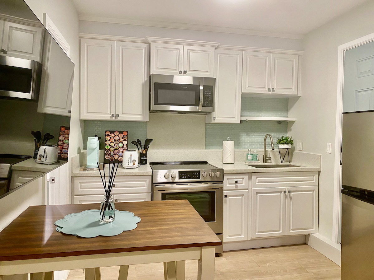 The kitchen area features modern stainless steel appliances, including a microwave and oven. Light-colored cabinets provide ample storage, while a compact dining table with a decorative centerpiece occupies the space. Soft lighting enhances the clean, fresh design of the kitchen.