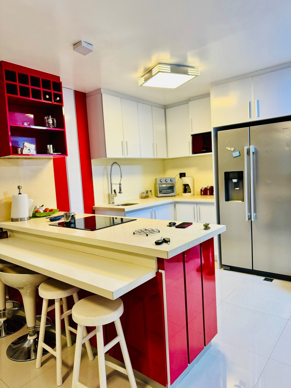 A modern kitchen features sleek white cabinetry paired with vibrant red accents. A central island with a cooktop is surrounded by three stool chairs. Stainless steel appliances, including a refrigerator and oven, complete the space, while the bright lighting enhances the overall ambiance.