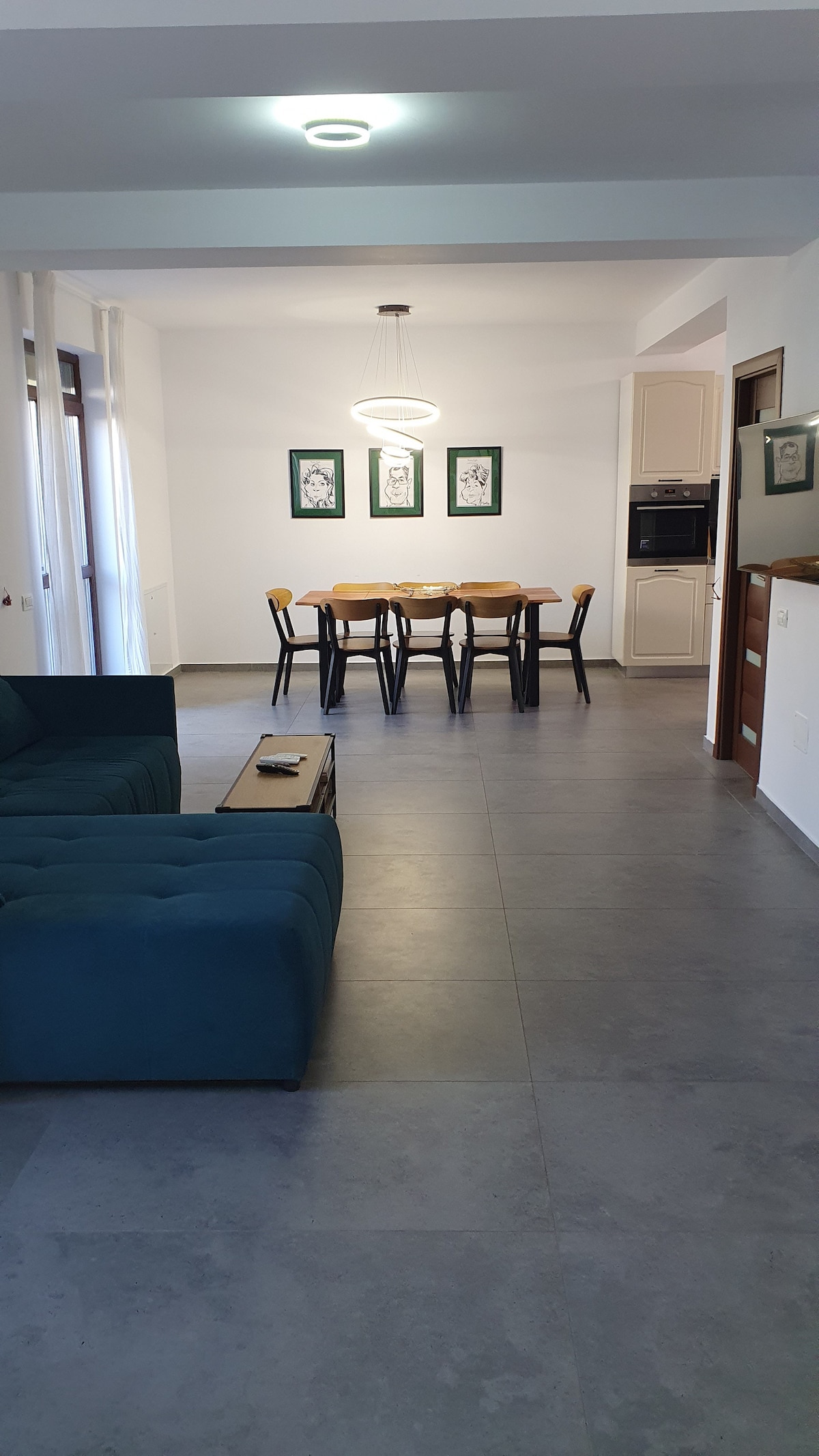 The living area features a spacious green couch facing a dining table set for six. Light-colored walls are complemented by framed artwork, while a modern light fixture hangs above the table. Sliding glass doors allow natural light to fill the space.