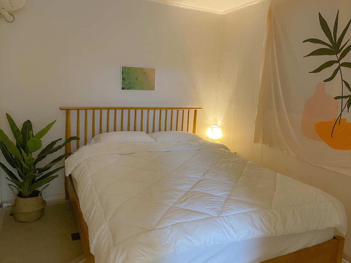 A serene bedroom features a spacious bed with a white comforter, complemented by a wooden frame. A bedside lamp provides soft lighting, while a green plant adds a touch of nature. A wall hanging with abstract shapes enhances the calming atmosphere.