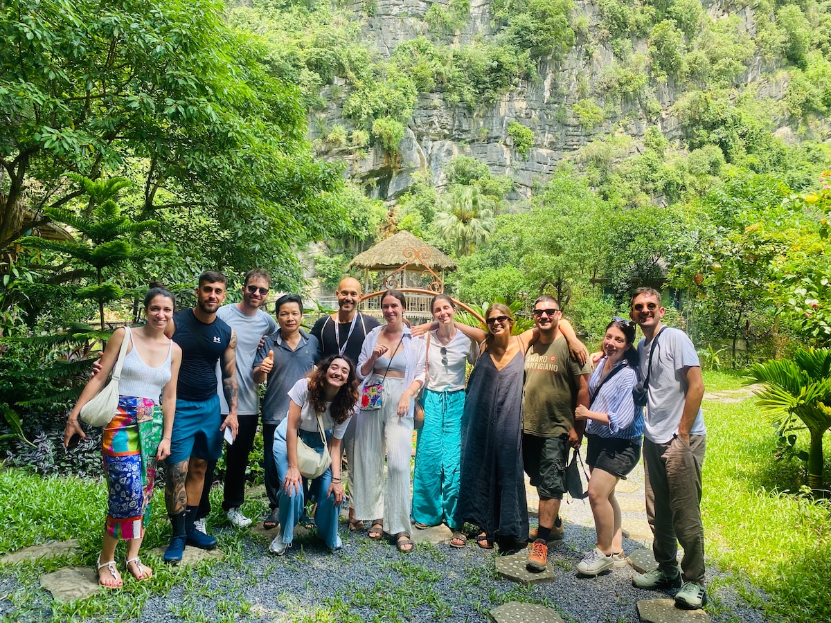 A group of twelve individuals stands together in a lush garden, surrounded by vibrant greenery and stone pathways. A wooden gazebo is visible in the background, nestled among the trees, contributing to the natural atmosphere of the area.