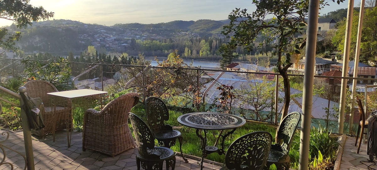 An outdoor terrace features a round metal table surrounded by four matching chairs, with woven armchairs nearby. The lush greenery and garden elements frame the space, offering views of the Rioni River and the surrounding hills, enhanced by soft morning light.
