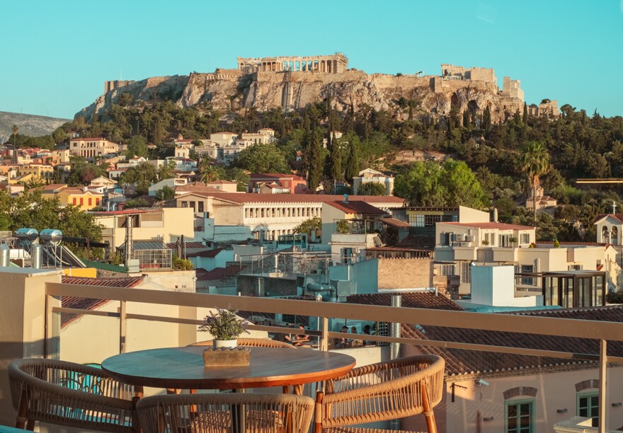 A private terrace offers a wooden dining table with four chairs, providing an unobstructed view of the Acropolis. The surrounding cityscape features varied architectural styles, with greenery interspersed among the buildings, all set against a clear blue sky.
