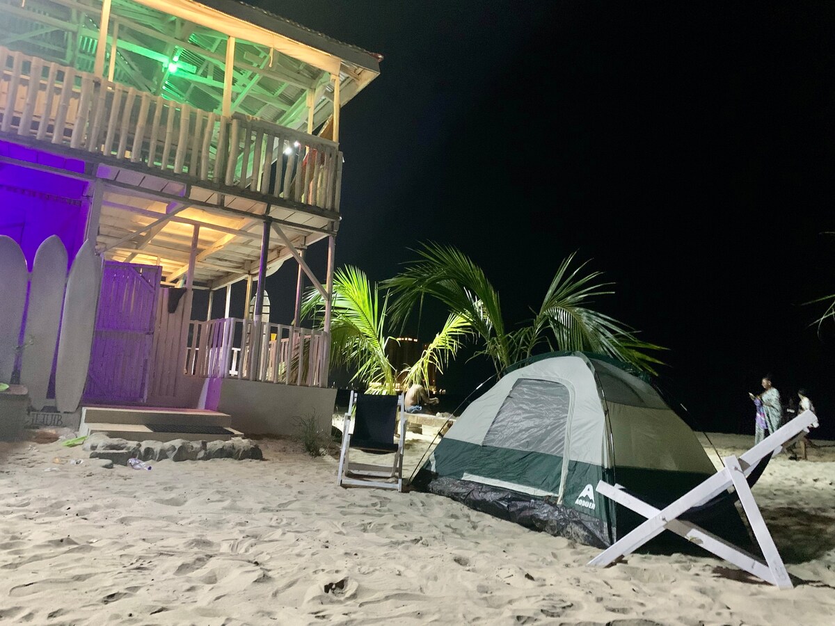 A tent is situated on the sandy beach, surrounded by palm trees. A two-story structure with wooden balconies stands nearby, illuminated with colored lights. A surfboard is positioned against the building, adding to the coastal ambiance. The night sky is clear, enhancing the serene beach setting.