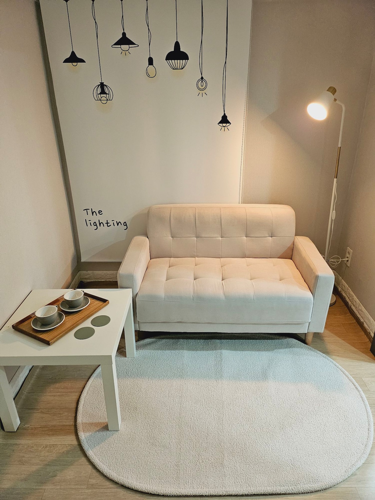A light-colored armchair is positioned next to a small table set with two cups. A soft rug lies beneath the furniture, and a floor lamp illuminates the space. The wall features playful lighting illustrations, contributing to a cozy ambiance.