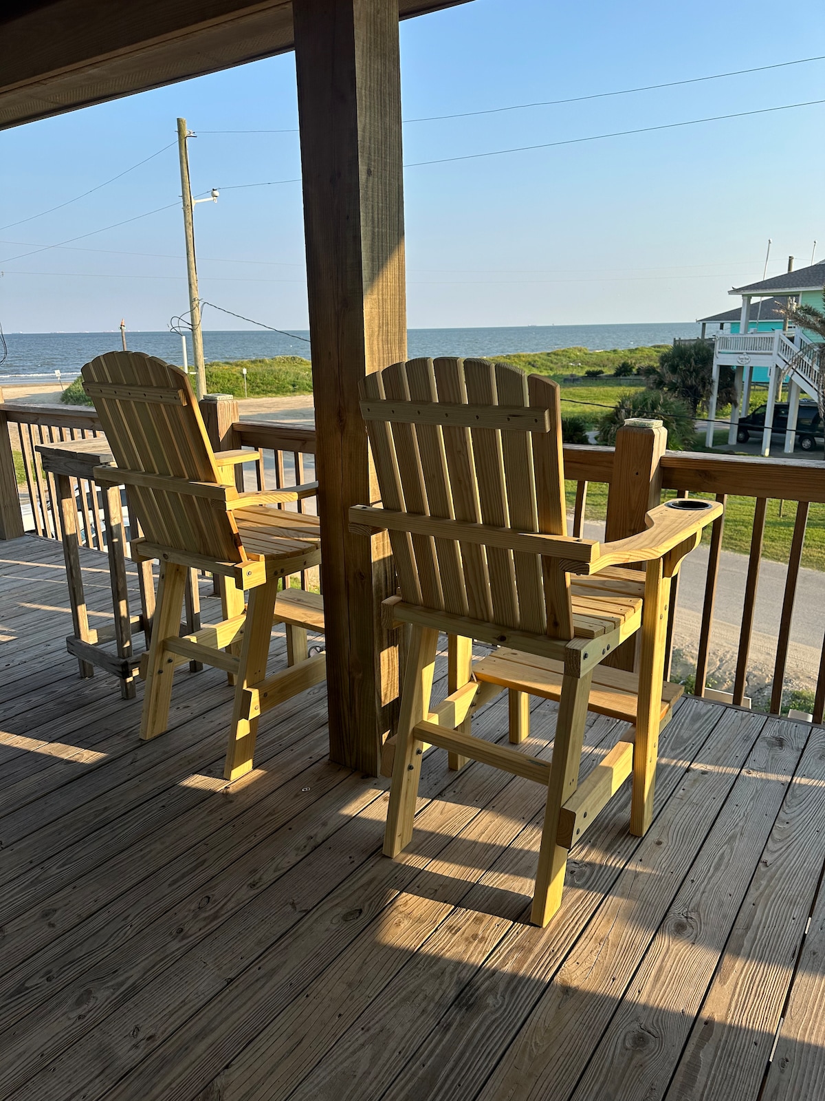 Two wooden high chairs are positioned on a spacious deck, overlooking a scenic view of the beach and the Gulf. The sun casts a warm glow, highlighting the natural wood finish. The surrounding landscape features grass and a distant beach house.