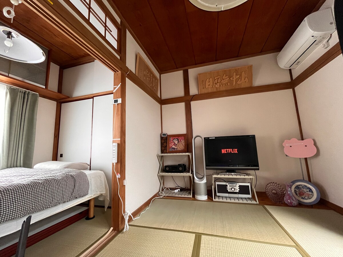 A tranquil bedroom features a double bed with soft bedding, a traditional tatami floor, and a television displaying a streaming service. Walls are adorned with simple wooden panels, and a window allows natural light to brighten the space.