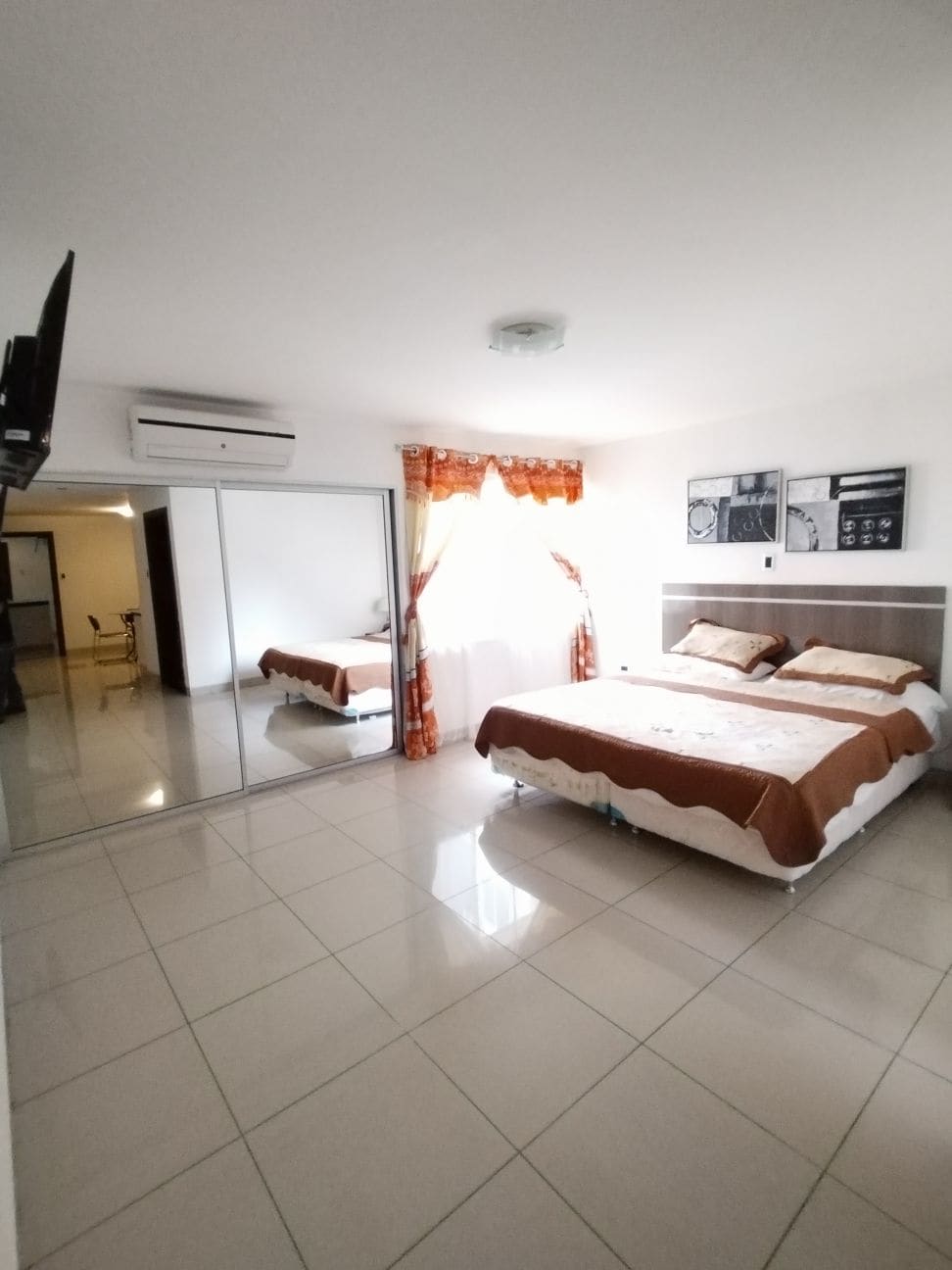 A modern bedroom features a spacious bed with neutral bedding and decorative pillows. A mirrored wardrobe reflects the light and ample space. Natural light filters through sheer curtains, adding brightness to the room, while air conditioning is mounted above for comfort.