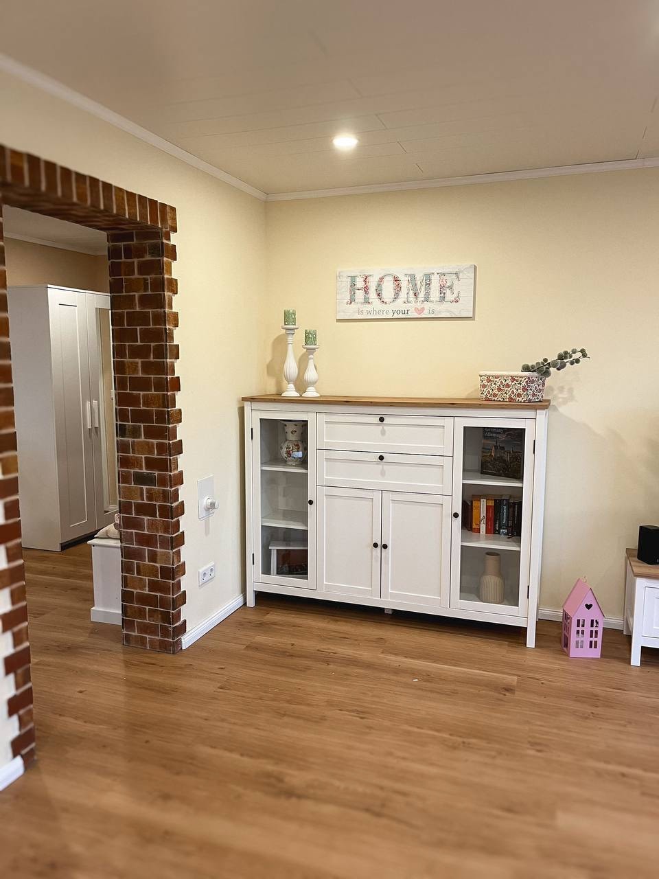 A welcoming living space displays a white cabinet featuring glass doors, showcasing decorative items and books. The wall is adorned with a framed sign reading 'HOME,' while warm wooden flooring complements the light-colored walls. An exposed brick archway adds character to the room.