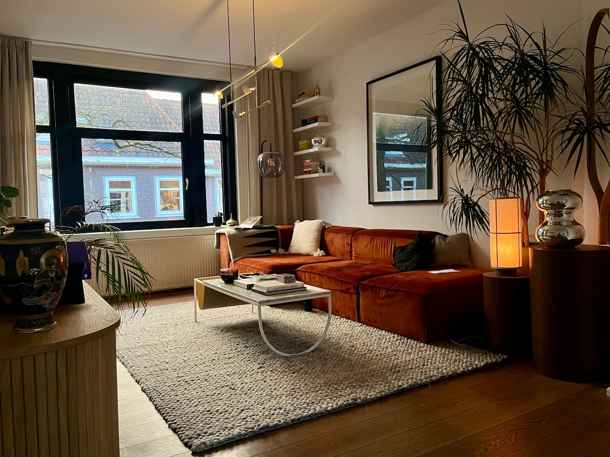 A well-appointed living area features a rich-colored sofa and an elegant coffee table positioned on a textured rug. Shelves display decorative items, while plants add a touch of greenery. Large windows provide ample natural light, enhancing the welcoming ambiance of the space.