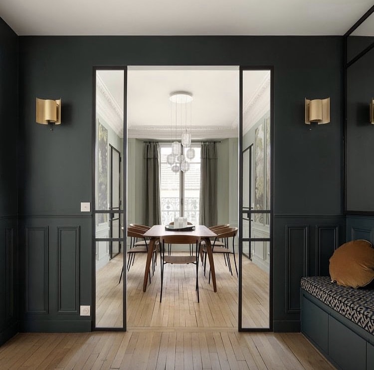 A dining area is framed by glass doors, featuring a long wooden table surrounded by mid-century style chairs. Elegant wall sconces and a modern chandelier provide tasteful lighting. Large windows with soft curtains allow natural light to fill the room, highlighting the rich green walls.