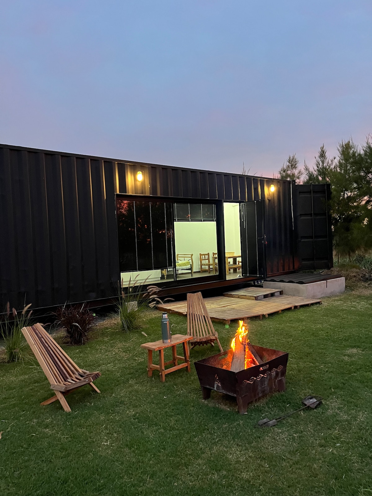 A modern container home is set against a twilight sky, featuring large glass doors that open to a wooden deck. In front, a fire pit surrounded by three wooden chairs provides a cozy gathering spot, with lush grass framing the scene.