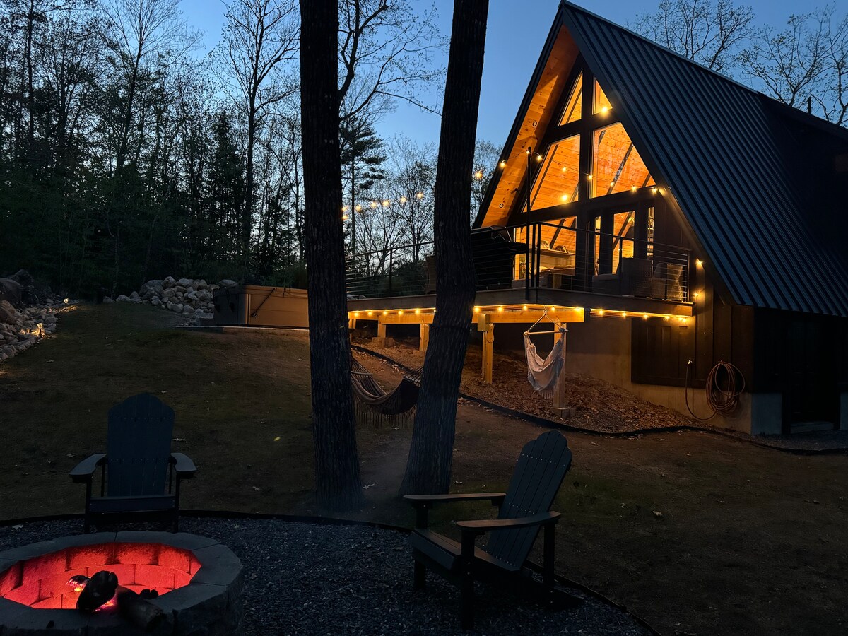 An A-frame retreat is illuminated at dusk, showcasing warm lights that accent the architectural features. In the foreground, a fire pit is surrounded by comfortable chairs, inviting cozy gatherings. Hammocks are seen hanging between trees, enhancing the serene outdoor environment.
