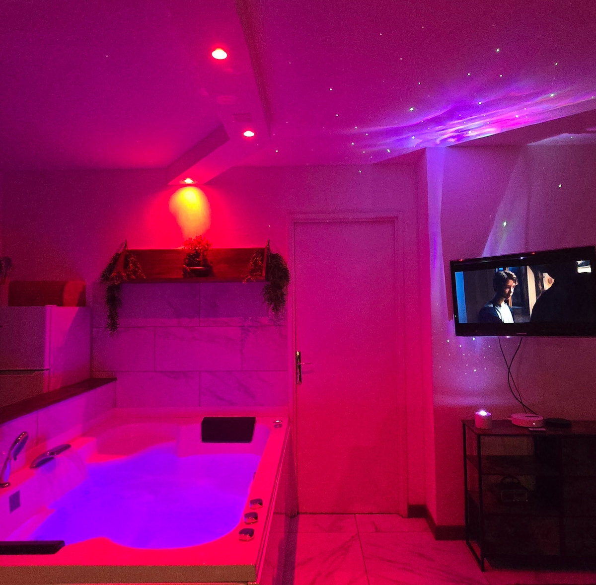 A spacious bathroom is equipped with a relaxing hot tub featuring LED lighting in soft hues. A television mounted on the wall displays programming, while decorative elements enhance the serene setting. Soft lights create a calming atmosphere, complemented by minimalist furnishings.