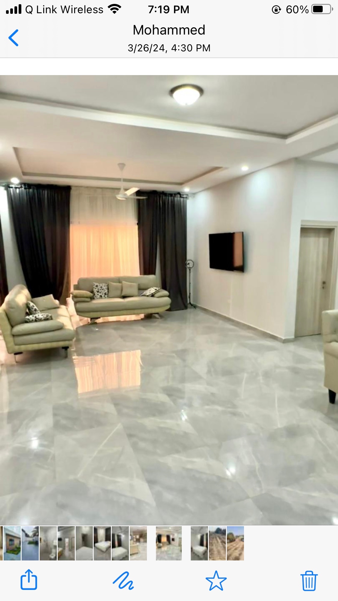 A spacious living area is illuminated by natural light filtering through large curtains. Light-colored sofas are arranged for comfortable seating, complemented by a flat-screen TV mounted on the wall. The tiled floor enhances the modern aesthetic of the room.