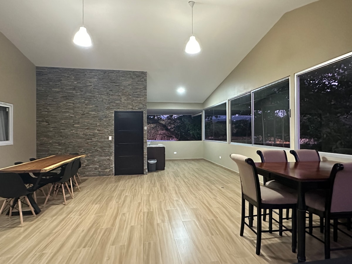 A spacious communal area is highlighted by large windows that provide ample natural light. A long dining table with black chairs is complemented by a round table with upholstered seating. Textured stone walls offer a unique backdrop, enhancing the room's modern aesthetic.