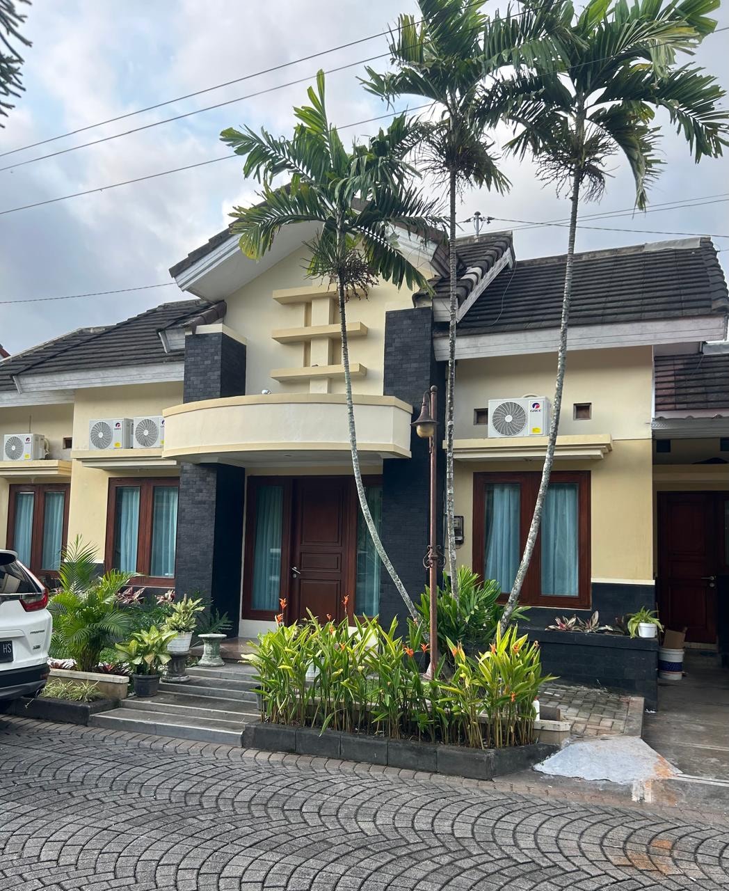 The exterior of the property is shown, featuring a two-story house with a welcoming façade. Large windows and a balcony are visible. Lush greenery surrounds the entrance, complemented by palm trees. Multiple air conditioning units are mounted on the walls, indicating a comfortable environment.
