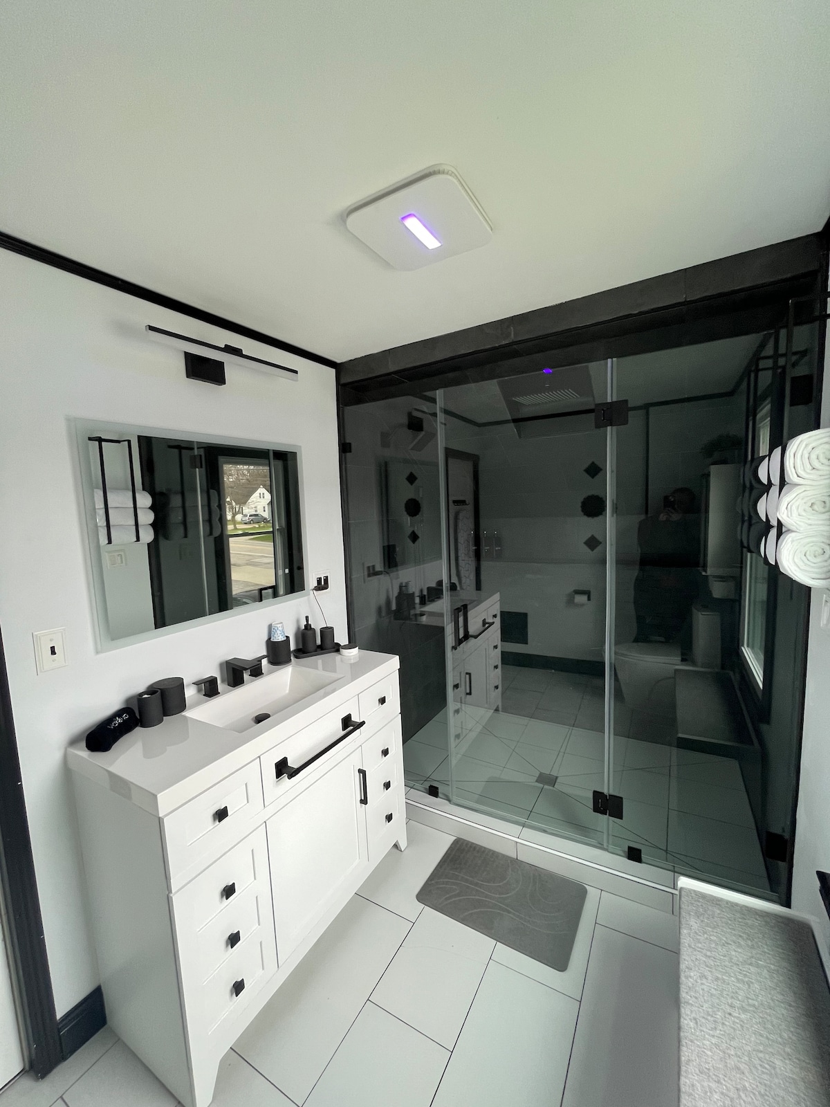 A modern bathroom features a glass shower enclosed by black-framed panels. A white vanity with multiple drawers provides storage, complemented by neatly arranged toiletries. Soft towels are rolled and displayed, enhancing the clean aesthetic. Natural light illuminates the space through a window.