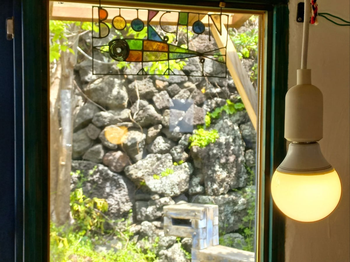 A window features a stained glass design reading 'Bookcan' above. Natural stone walls display various textures and colors, while daylight illuminates the area. A minimalist pendant light hangs from the ceiling, providing warm lighting to the space.