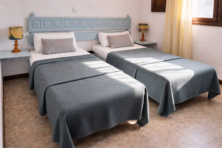 Dhomë dyshe e ndritshme dhe e ajrosur me dy krevate të rehatshme tekë, tone të buta neutrale dhe thekse elegante blu. Ka dritë natyrale të diellit, abazhurë pranë krevatit dhe një ambient të qetë e tërheqës, ideal për një qëndrim çlodhës dhe relaksues.