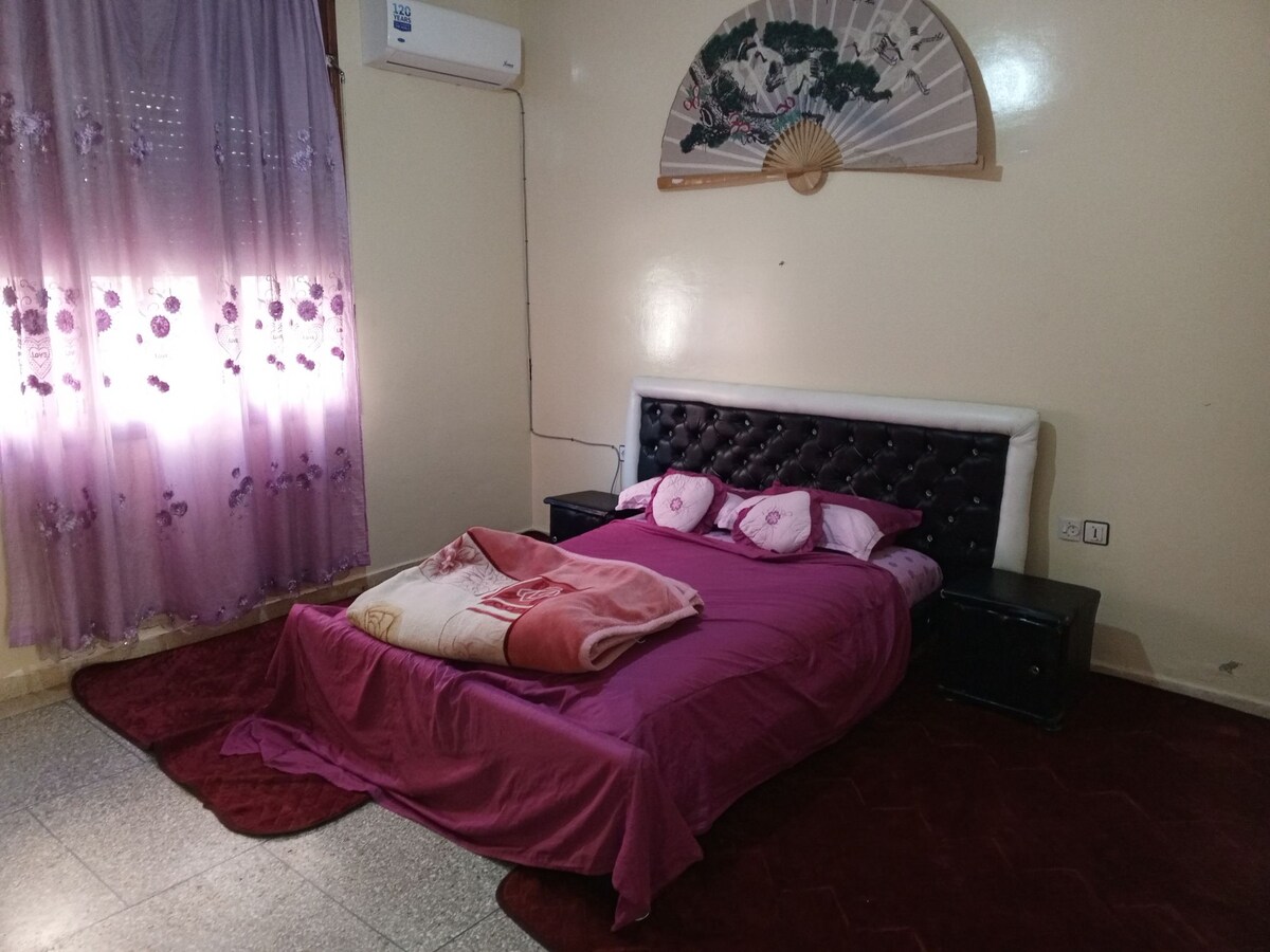 A bedroom is equipped with a spacious bed draped in a purple coverlet. Two nightstands sit on either side of the bed, and a fan is visible on the wall above. Sheer purple curtains allow soft light to filter into the room.