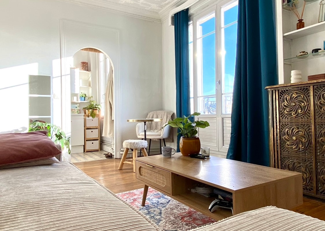 The living area features a wooden coffee table with a potted plant at its center. A comfortable chair is positioned nearby, while large windows allow natural light to fill the space. Light blue curtains frame the windows, contributing to a serene ambiance.