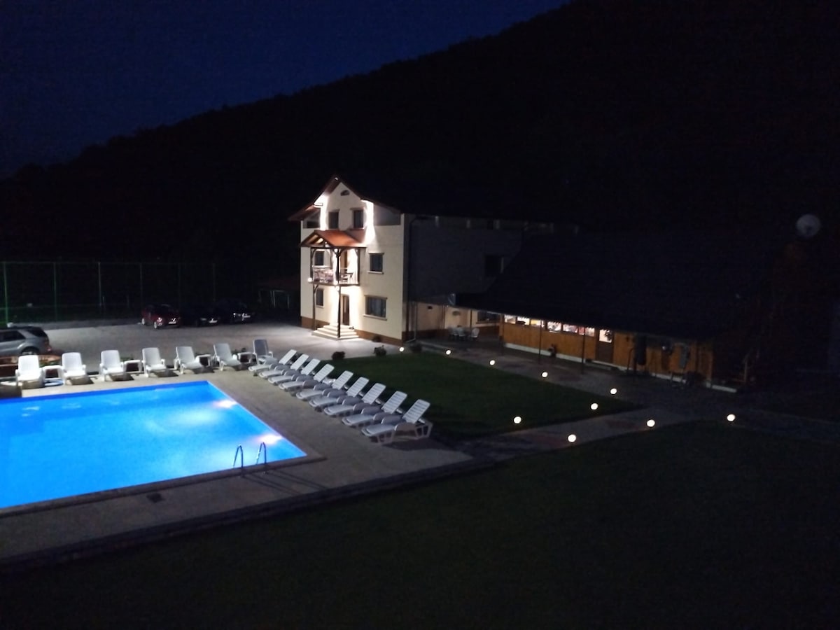 A well-lit outdoor area is showcased at night, featuring a swimming pool with blue water. Surrounding the pool are multiple lounge chairs arranged neatly. An adjacent building is partially illuminated, indicating a welcoming space for gatherings.