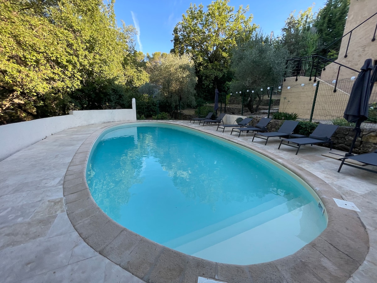A serene outdoor pool is surrounded by a terrace featuring sun loungers. The pool reflects the clear sky and greenery surrounding the area, offering a tranquil space for relaxation. Olive trees and shrubs provide a natural backdrop, enhancing the peaceful setting.
