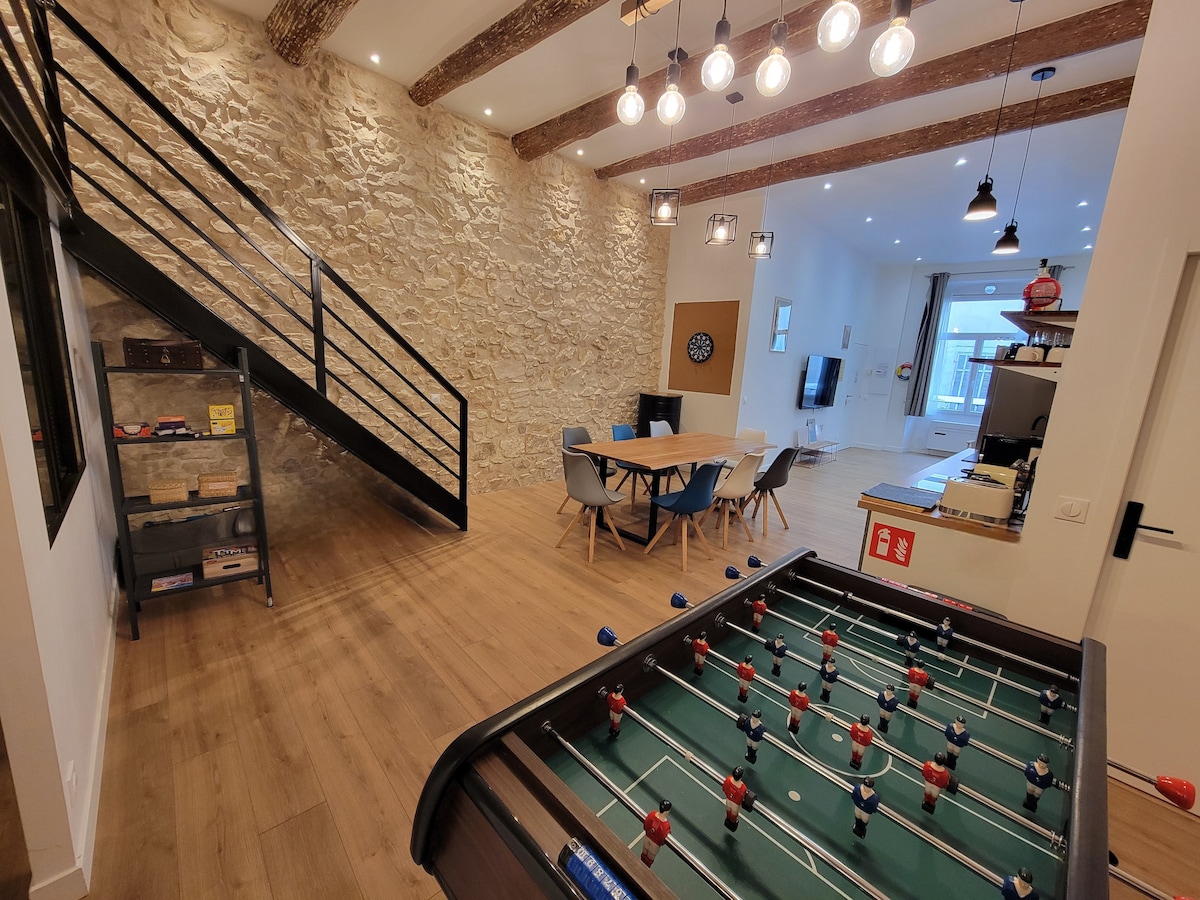 A spacious living area features a foosball table and a dining table surrounded by modern chairs. Exposed stone walls contrast with reclaimed wood beams overhead. An open staircase leads to the mezzanine, while a large window brings natural light into the room.
