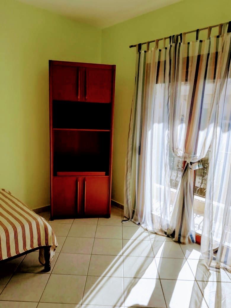 A room features light green walls and a spacious bed with striped bedding. A wooden shelving unit occupies one corner, while bright natural light filters through sheer curtains, illuminating the tiled floor.