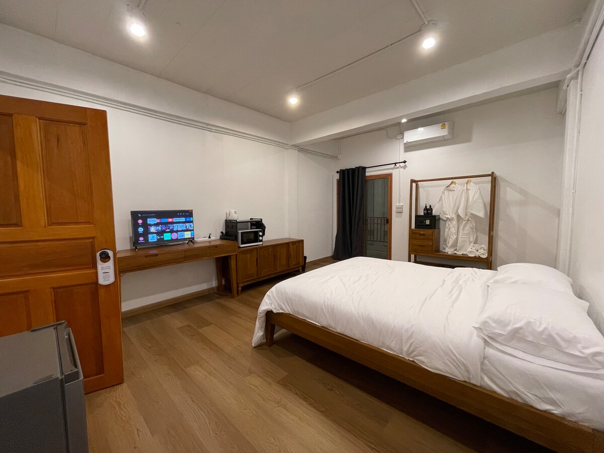 A minimalist bedroom features a wooden bed with a neatly arranged white duvet. A wooden desk holds a television and a small microwave, while a chest of drawers displays hanging garments. The space is brightened by artificial lighting and includes a window dressed with dark curtains.