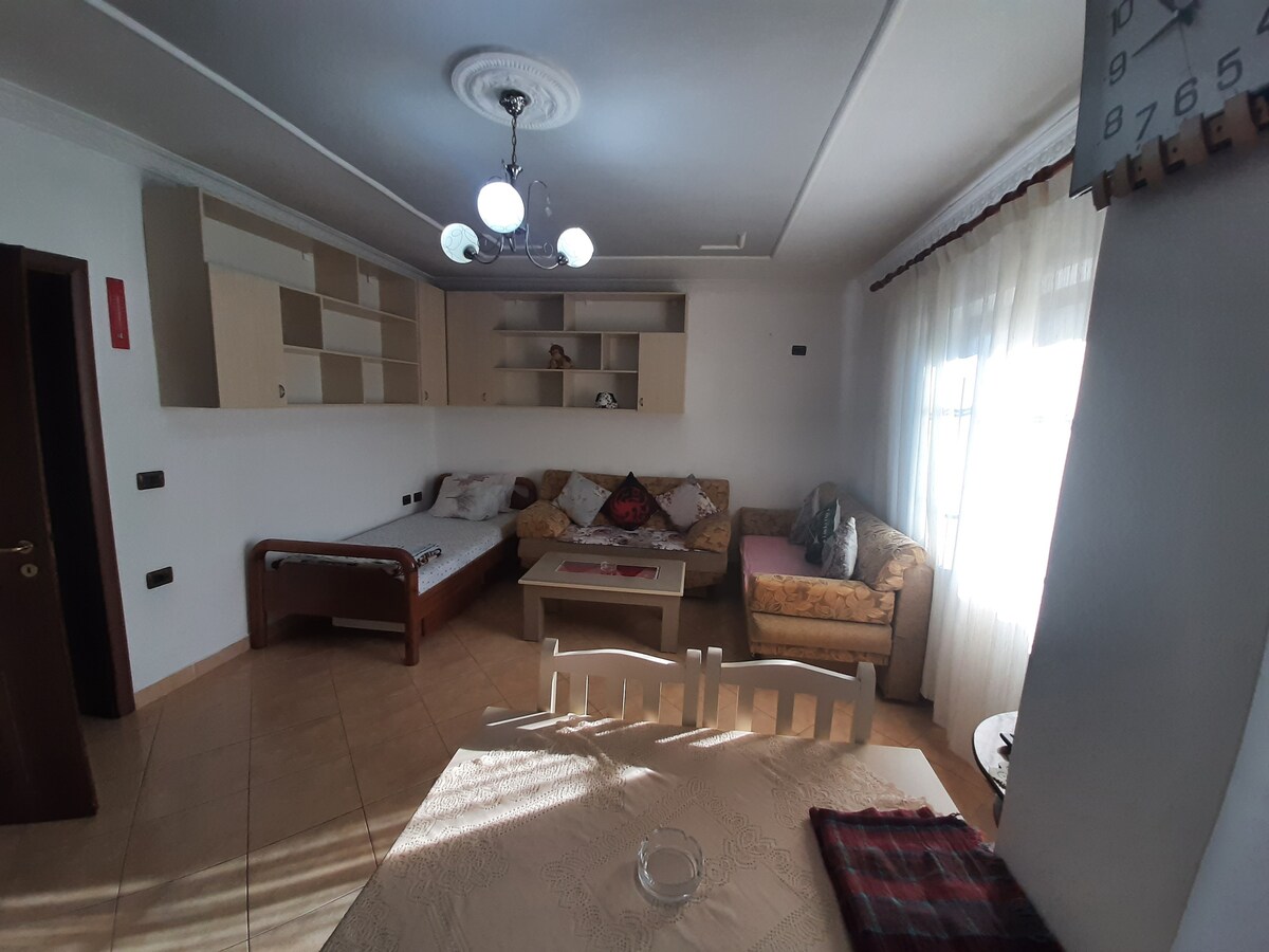 A bright living area features a spacious seating arrangement with a mix of couches and chairs. Natural light filters through the window, highlighting the light-colored walls and tiled floor. A dining table is situated in the foreground, with a decorative runner on its surface.