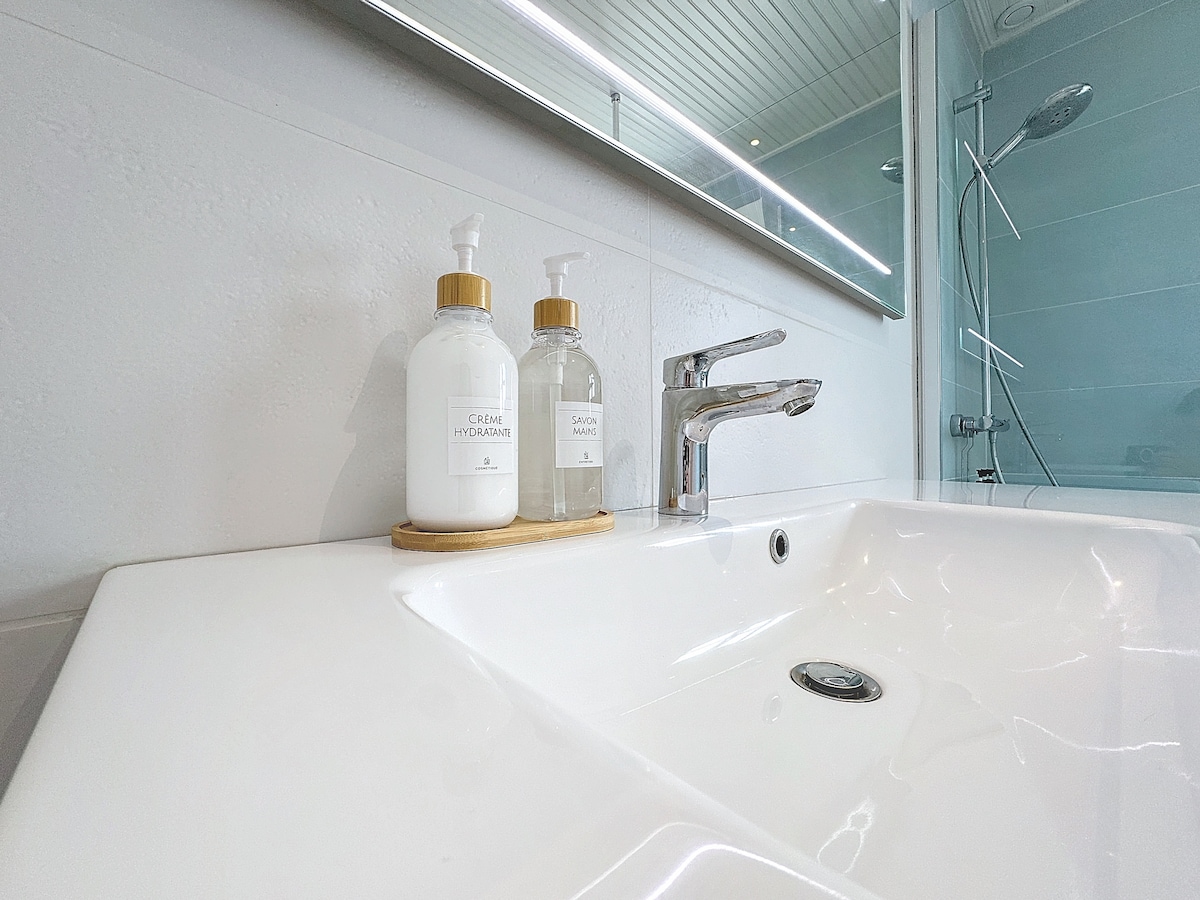 The modern bathroom features a sleek white sink with a polished faucet. On the countertop, two elegant soap dispensers in glass bottles are placed on a wooden tray, alongside a mirror that reflects the light from the room's fixtures.