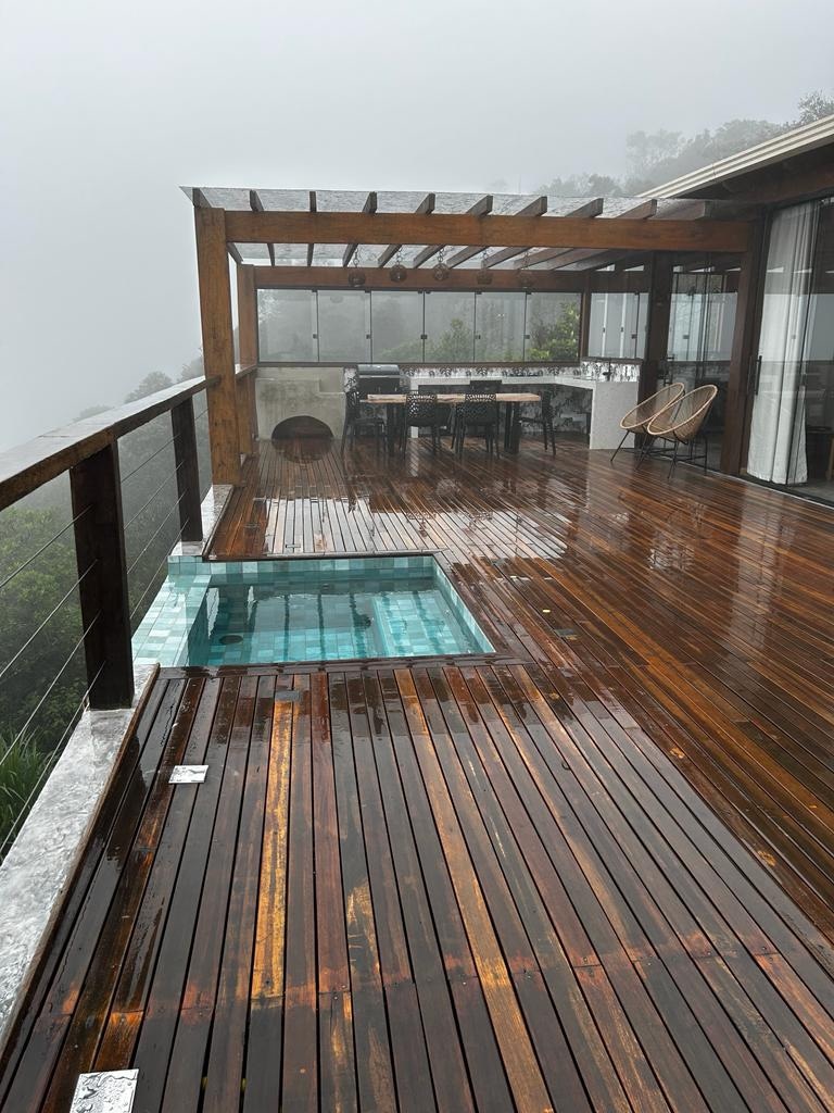 A spacious wooden deck is depicted, featuring a small pool with turquoise tiles. A dining area is visible with a large table and chairs, while two lounge chairs are positioned in a corner. The surrounding mist creates a serene atmosphere.