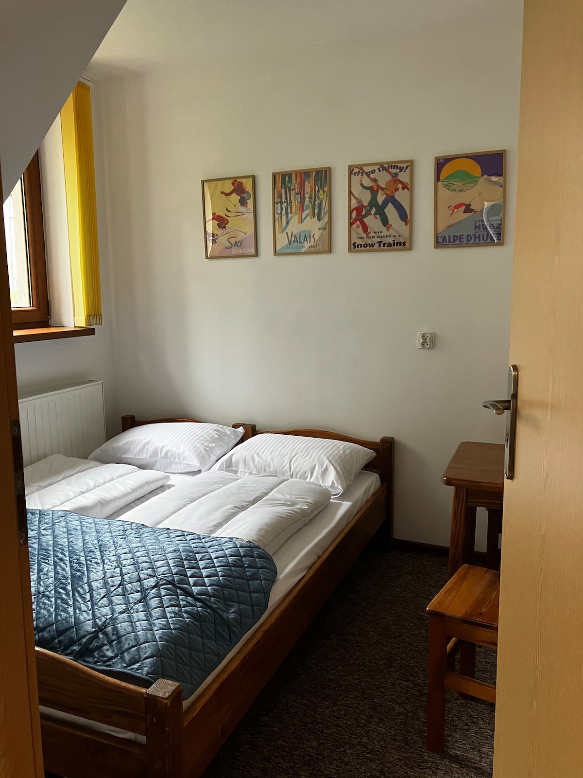 A cozy bedroom is visible through an open door, featuring a wooden bed with soft bedding and decorative pillows. Wall-mounted vintage posters add character, while a small wooden table and chair complement the space. Natural light brightens the room from a nearby window.