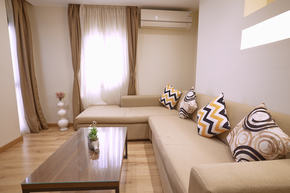 A comfortable living area features a light-colored sofa adorned with patterned throw pillows. A wooden coffee table sits in front, while large windows are framed by soft curtains that allow natural light to fill the space. A small decorative plant adds a touch of greenery.