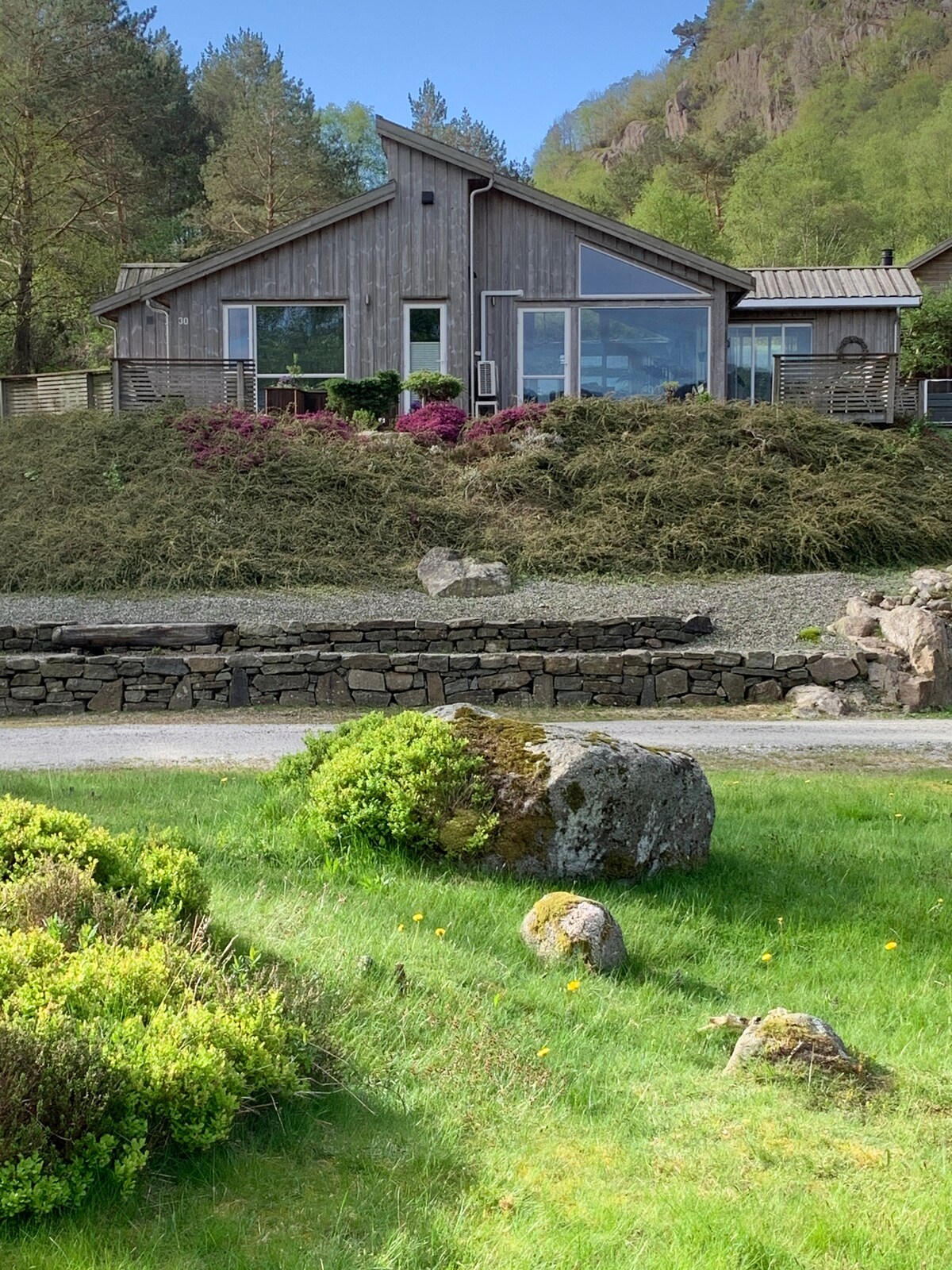 A modern wooden cabin features large glass panels that provide views of the surrounding greenery. The exterior is complemented by vibrant landscaping, including flowering bushes and carefully arranged stones, set against a backdrop of gently sloping hills.