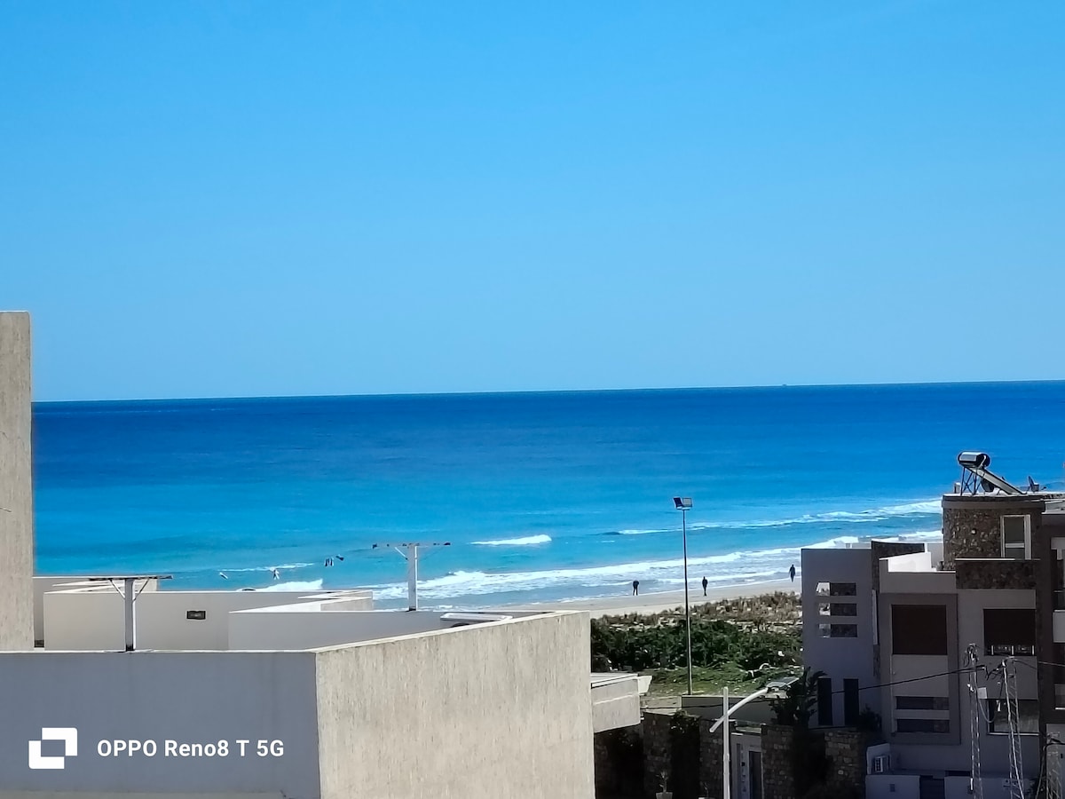An expansive view of the ocean is visible, showcasing vibrant shades of blue and gentle waves. The horizon blends seamlessly with the sky, creating a tranquil atmosphere. In the foreground, buildings are positioned along the shoreline, with people enjoying the beach.